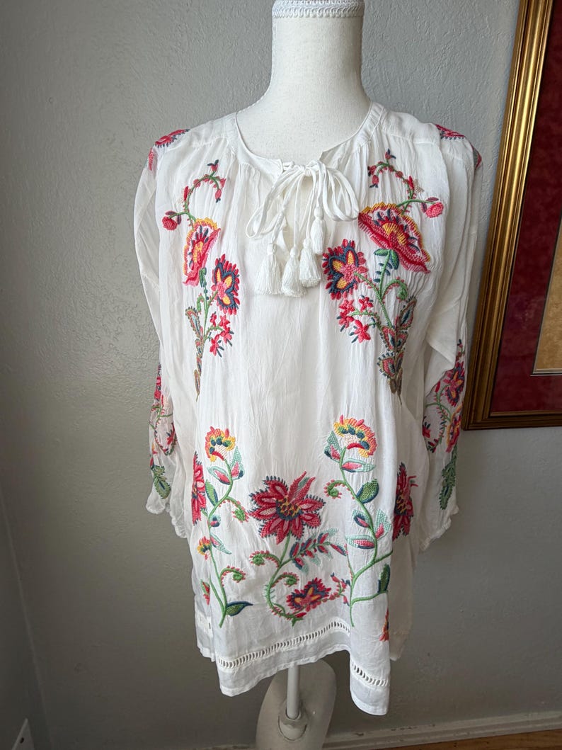 May include: White tunic top with colorful floral embroidery. The long-sleeved top features red, pink, yellow, and green embroidered flowers and leaves. It has a round neckline with a tie and tassel detail.
