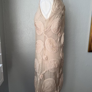 May include: A sleeveless, beige dress with a V-neckline. The dress is adorned with large, textured, rose-shaped appliqu&eacute;s and delicate, flowing vines. The dress is displayed on a mannequin.