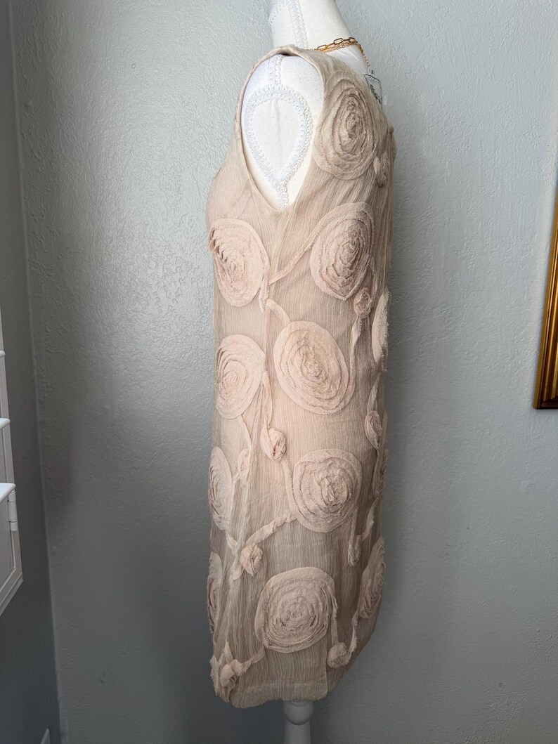 May include: A sleeveless, beige dress with a V-neckline. The dress is adorned with large, textured, rose-shaped appliqu&eacute;s and delicate, flowing vines. The dress is displayed on a mannequin.