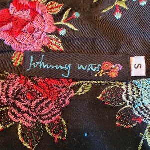 May include: Close-up of a black garment with vibrant floral embroidery in red, pink, blue, and yellow. The label reads "Johnny Was" in turquoise script, with a small floral design. A size "S" tag is visible.