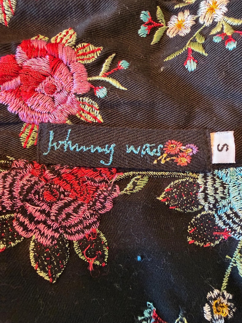 May include: Close-up of a black garment with vibrant floral embroidery in red, pink, blue, and yellow. The label reads "Johnny Was" in turquoise script, with a small floral design. A size "S" tag is visible.