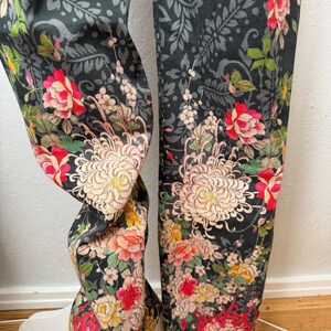 Puede incluir: Un pantal&oacute;n con fondo gris oscuro y un patr&oacute;n floral denso e intrincado. El dise&ntilde;o incluye grandes crisantemos de color rosa p&aacute;lido, rosas de color rojo vibrante y flores m&aacute;s peque&ntilde;as amarillas y blancas sobre un estampado de follaje gris.