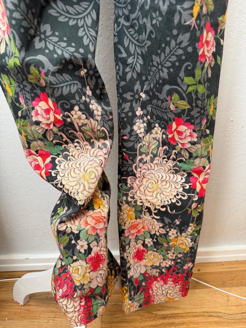 Puede incluir: Un pantal&oacute;n con fondo gris oscuro y un patr&oacute;n floral denso e intrincado. El dise&ntilde;o incluye grandes crisantemos de color rosa p&aacute;lido, rosas de color rojo vibrante y flores m&aacute;s peque&ntilde;as amarillas y blancas sobre un estampado de follaje gris.
