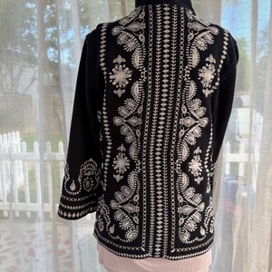 K&ouml;nnte beinhalten: Schwarze Jacke mit aufwendiger wei&szlig;er Stickerei auf R&uuml;cken und &Auml;rmeln. Die Stickerei zeigt florale und Paisley-Muster. Die Jacke hat einen hohen Kragen und lange &Auml;rmel. Pr&auml;sentiert auf einer Schaufensterpuppe.