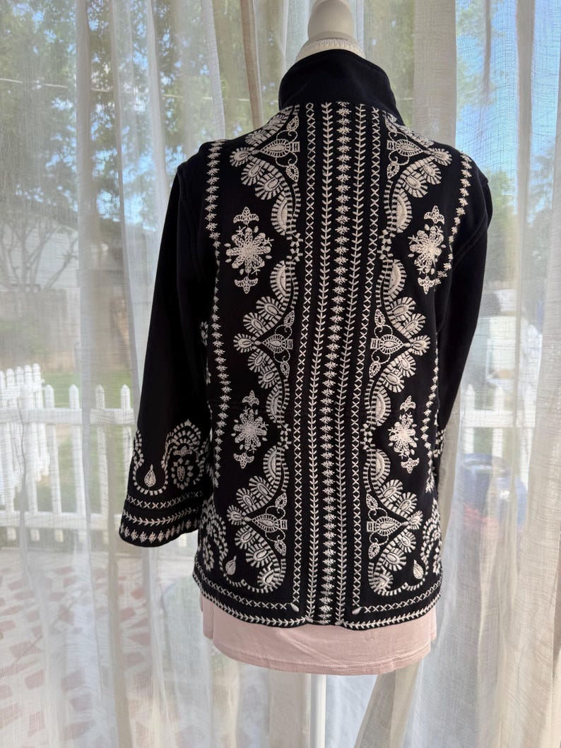 K&ouml;nnte beinhalten: Schwarze Jacke mit aufwendiger wei&szlig;er Stickerei auf R&uuml;cken und &Auml;rmeln. Die Stickerei zeigt florale und Paisley-Muster. Die Jacke hat einen hohen Kragen und lange &Auml;rmel. Pr&auml;sentiert auf einer Schaufensterpuppe.