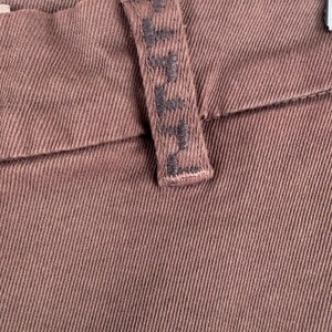 May include: Close-up of brown denim trousers. The trousers feature a textured fabric with a visible seam and a belt loop. The belt loop has embroidered text. The overall colour is a muted brown.