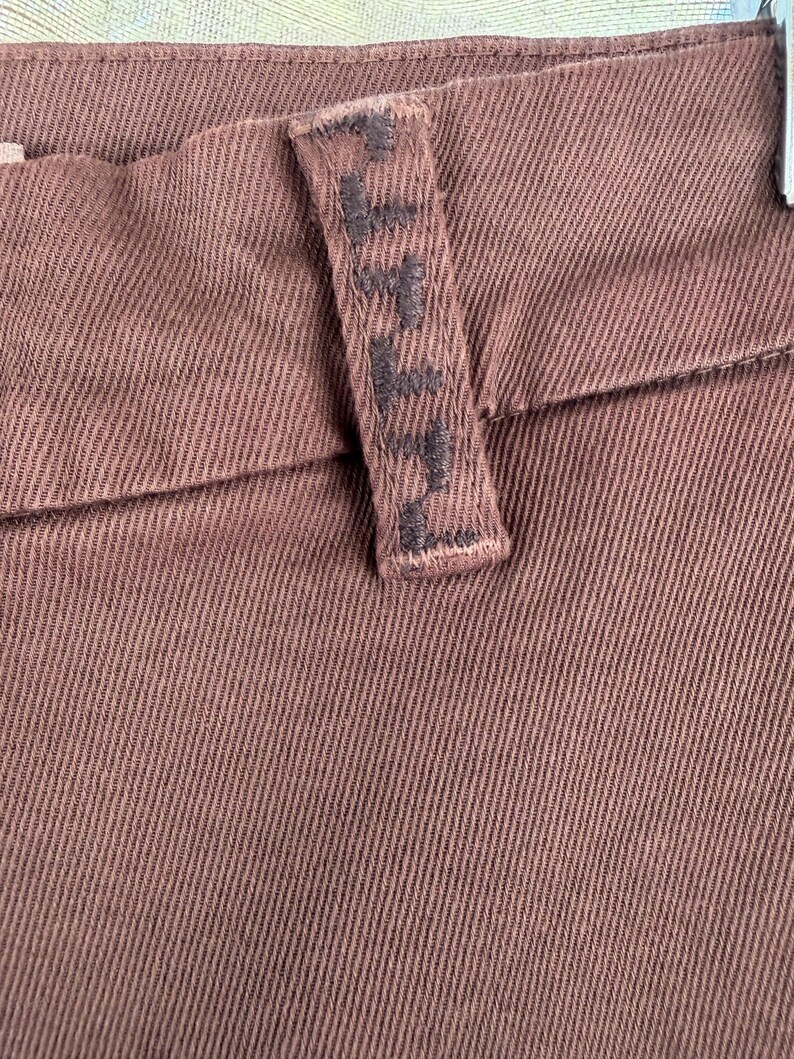 May include: Close-up of brown denim trousers. The trousers feature a textured fabric with a visible seam and a belt loop. The belt loop has embroidered text. The overall colour is a muted brown.