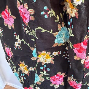 May include: Black jacket with colorful floral embroidery. The embroidery features pink, red, yellow, and blue flowers with green leaves. The jacket is made of a dark fabric and has a detailed, textured design.