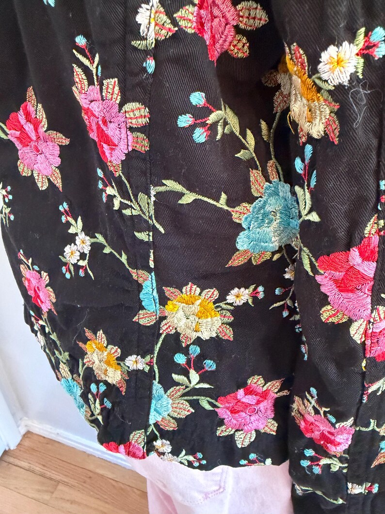 May include: Black jacket with colorful floral embroidery. The embroidery features pink, red, yellow, and blue flowers with green leaves. The jacket is made of a dark fabric and has a detailed, textured design.