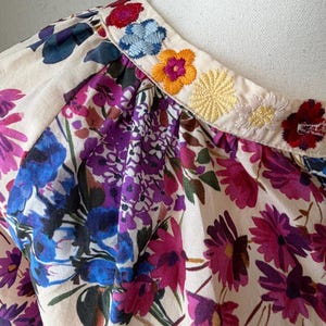 May include: Close-up of a floral print top with a cream-colored background. The top features a vibrant pattern of purple, pink, and blue flowers. The neckline is adorned with embroidered flowers in various colors, including red, blue, and yellow.