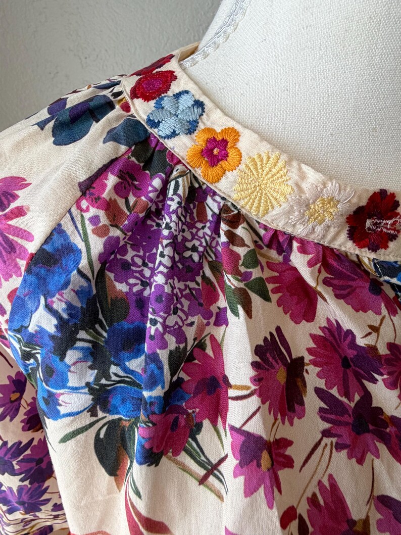 May include: Close-up of a floral print top with a cream-colored background. The top features a vibrant pattern of purple, pink, and blue flowers. The neckline is adorned with embroidered flowers in various colors, including red, blue, and yellow.