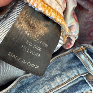 Op de afbeelding: Close-up van een zwart kledinglabel met witte tekst: "94% Katoen, 5% T400, 1% Lycra, Made in China." Het label is bevestigd aan een blauwe spijkerbroek met zichtbare stiksels en een knoop.