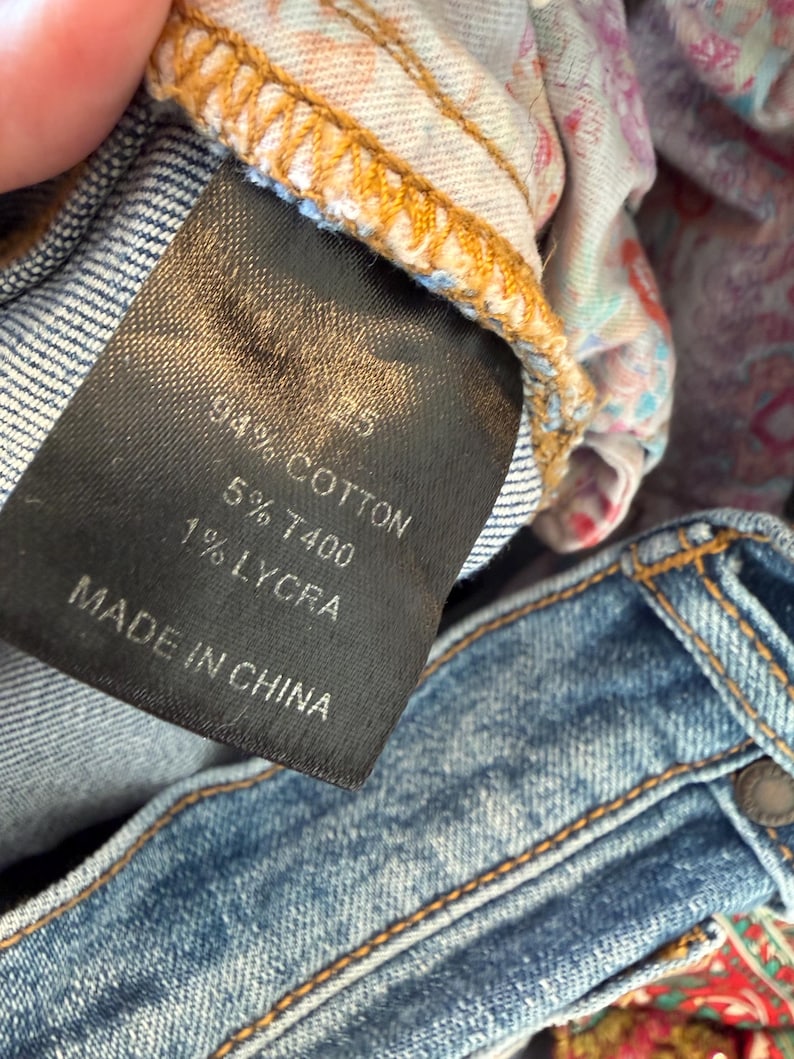 Op de afbeelding: Close-up van een zwart kledinglabel met witte tekst: "94% Katoen, 5% T400, 1% Lycra, Made in China." Het label is bevestigd aan een blauwe spijkerbroek met zichtbare stiksels en een knoop.