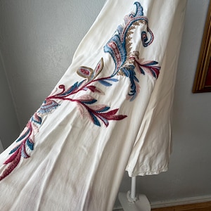 May include: Off-white dress with intricate embroidered floral patterns in shades of blue, red, and green. The embroidery is detailed and covers a portion of the dress, creating a decorative design.