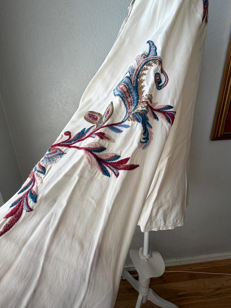 May include: Off-white dress with intricate embroidered floral patterns in shades of blue, red, and green. The embroidery is detailed and covers a portion of the dress, creating a decorative design.
