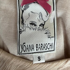 May include: Close-up of a woven label with the text "YOANA BARASCHI" in black, a portrait of a woman, and a small pink bow. The label is attached to a light beige fabric. The size "S" is printed on the label.