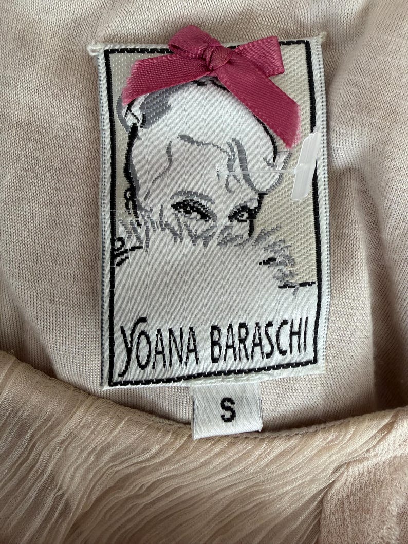 May include: Close-up of a woven label with the text "YOANA BARASCHI" in black, a portrait of a woman, and a small pink bow. The label is attached to a light beige fabric. The size "S" is printed on the label.
