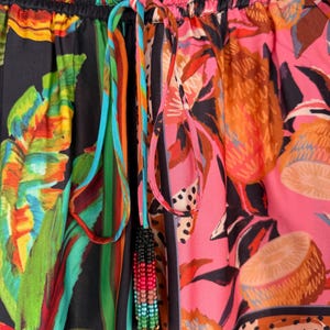 Puede incluir: Primer plano de pantalones estampados coloridos con cord&oacute;n. La tela presenta dise&ntilde;os florales y abstractos en tonos rosa, verde, naranja y negro. Una borla de cuentas cuelga del cord&oacute;n.