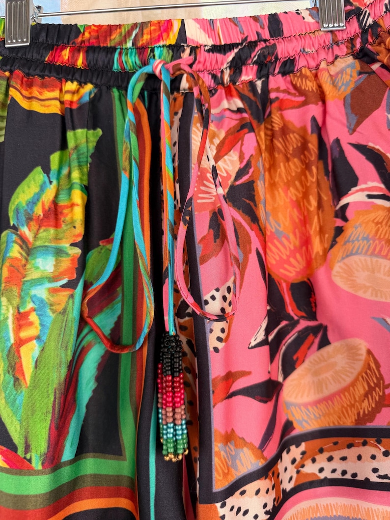 Puede incluir: Primer plano de pantalones estampados coloridos con cord&oacute;n. La tela presenta dise&ntilde;os florales y abstractos en tonos rosa, verde, naranja y negro. Una borla de cuentas cuelga del cord&oacute;n.