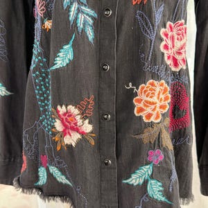 以下が含まれることがあります： カラフルな花の刺繍が施された、濃いグレーのボタンダウンシャツ。 刺繍には、オレンジ、ピンク、ターコイズの花と葉が使われています。 シャツは裾がほつれており、長袖です。