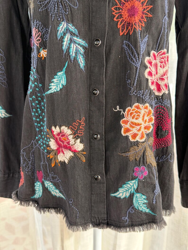 以下が含まれることがあります： カラフルな花の刺繍が施された、濃いグレーのボタンダウンシャツ。 刺繍には、オレンジ、ピンク、ターコイズの花と葉が使われています。 シャツは裾がほつれており、長袖です。