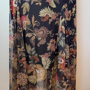 May include: A long, flowing black dress with a vibrant floral pattern. The design features intricate paisley and botanical elements in shades of gold, red, and green. The dress has a sheer overlay, adding a delicate touch to the overall design.