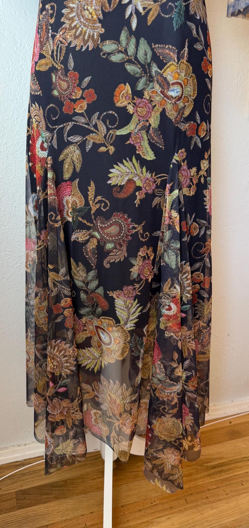 May include: A long, flowing black dress with a vibrant floral pattern. The design features intricate paisley and botanical elements in shades of gold, red, and green. The dress has a sheer overlay, adding a delicate touch to the overall design.