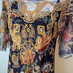 May include: Black dress with a floral pattern and gold embroidery. The dress has short sleeves and a sheer overlay. A gold necklace is draped over the dress.