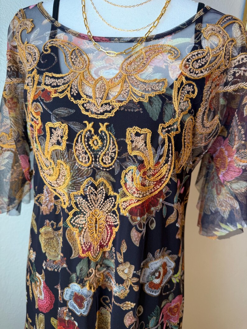 May include: Black dress with a floral pattern and gold embroidery. The dress has short sleeves and a sheer overlay. A gold necklace is draped over the dress.