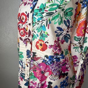 May include: A cream-colored button-down shirt with a vibrant floral pattern. The design features large, colorful flowers in shades of red, pink, blue, and orange, set against a backdrop of green leaves. The shirt has a classic collar and long sleeves.