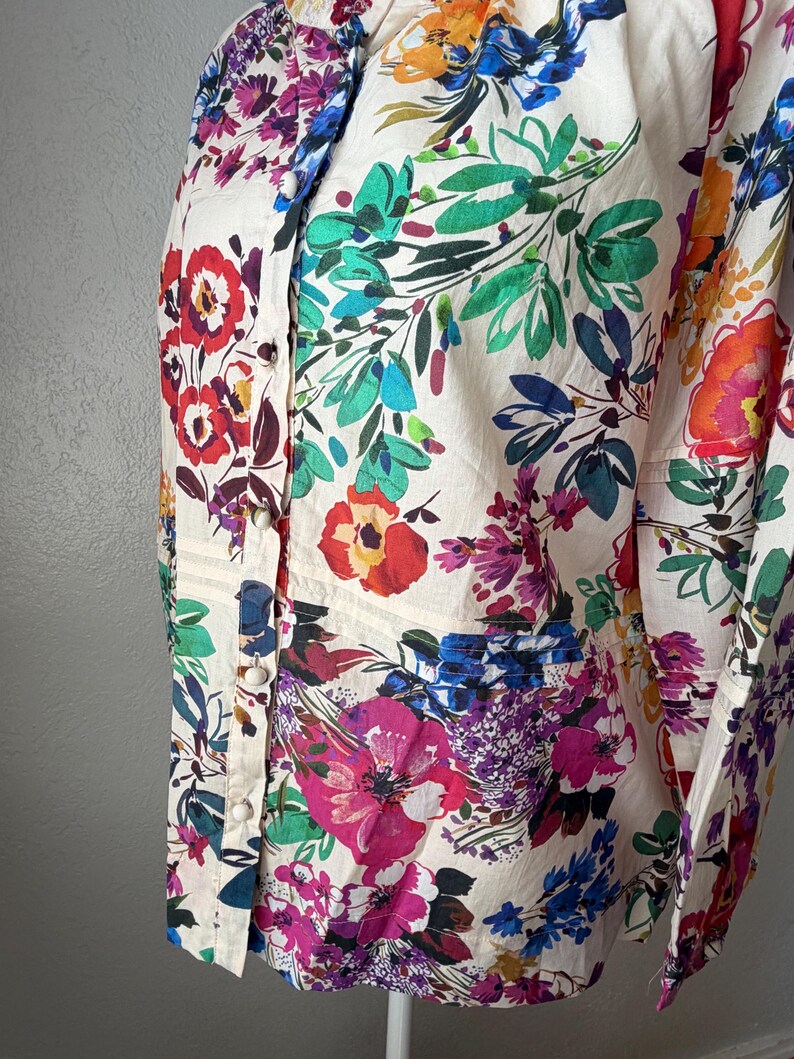 May include: A cream-colored button-down shirt with a vibrant floral pattern. The design features large, colorful flowers in shades of red, pink, blue, and orange, set against a backdrop of green leaves. The shirt has a classic collar and long sleeves.