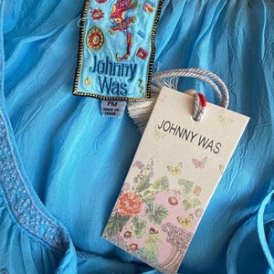 Puede incluir: Una blusa azul claro con detalles bordados y una etiqueta que dice "JOHNNY WAS". La etiqueta presenta ilustraciones florales y de mariposas. Tambi&eacute;n es visible una etiqueta m&aacute;s peque&ntilde;a con un dise&ntilde;o de lagarto y el nombre de la marca.