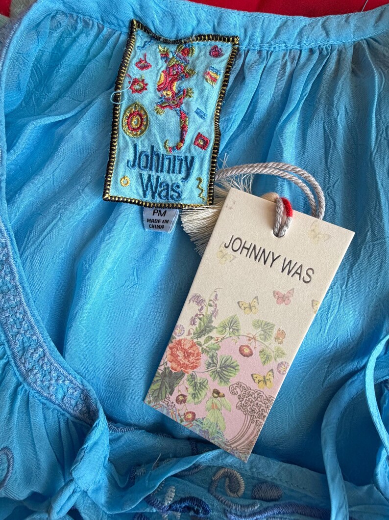 Puede incluir: Una blusa azul claro con detalles bordados y una etiqueta que dice "JOHNNY WAS". La etiqueta presenta ilustraciones florales y de mariposas. Tambi&eacute;n es visible una etiqueta m&aacute;s peque&ntilde;a con un dise&ntilde;o de lagarto y el nombre de la marca.