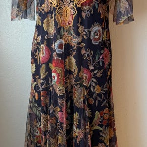 May include: A long, dark dress with a floral pattern. The dress features a sheer overlay with short sleeves and a gold-colored embroidered design around the neckline. The dress is displayed on a mannequin.