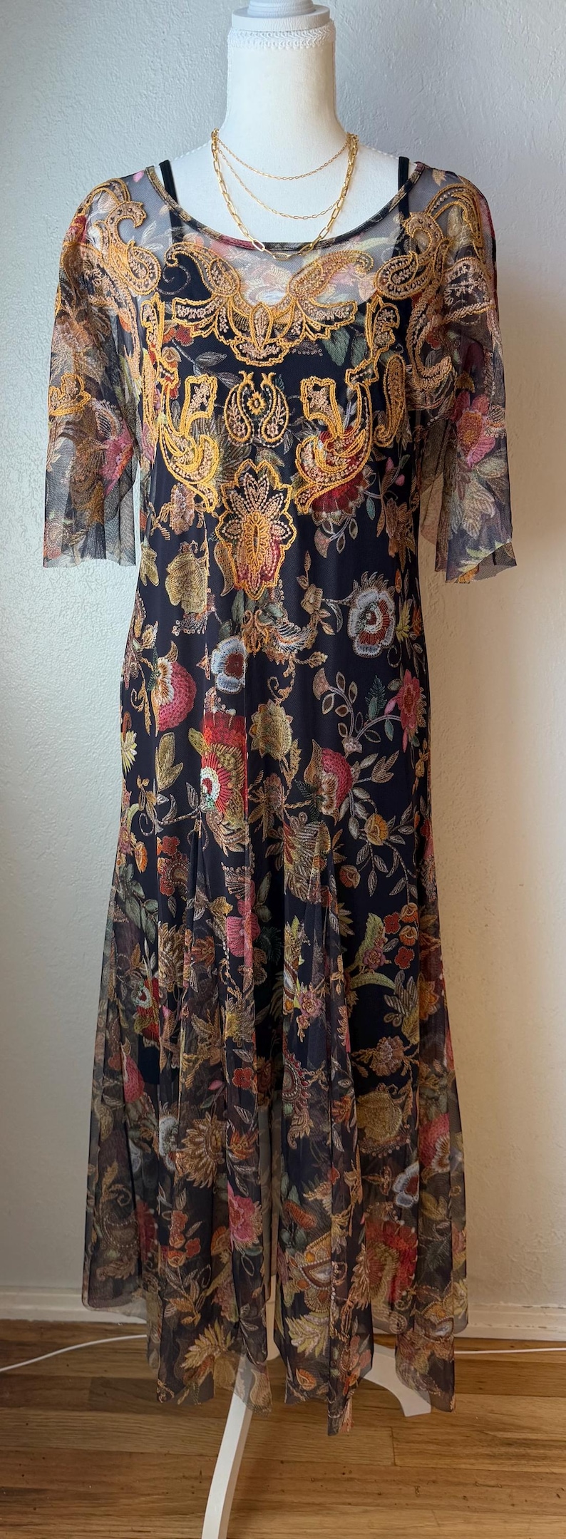 May include: A long, dark dress with a floral pattern. The dress features a sheer overlay with short sleeves and a gold-colored embroidered design around the neckline. The dress is displayed on a mannequin.