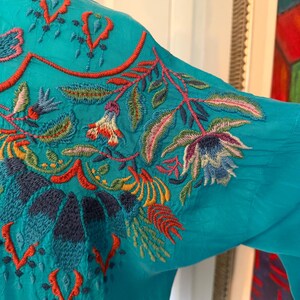 May include: Teal-colored blouse with intricate floral embroidery in shades of red, orange, blue, green, and yellow. The embroidery is concentrated on the upper back and shoulder area, creating a vibrant and detailed design. The blouse has long sleeves.