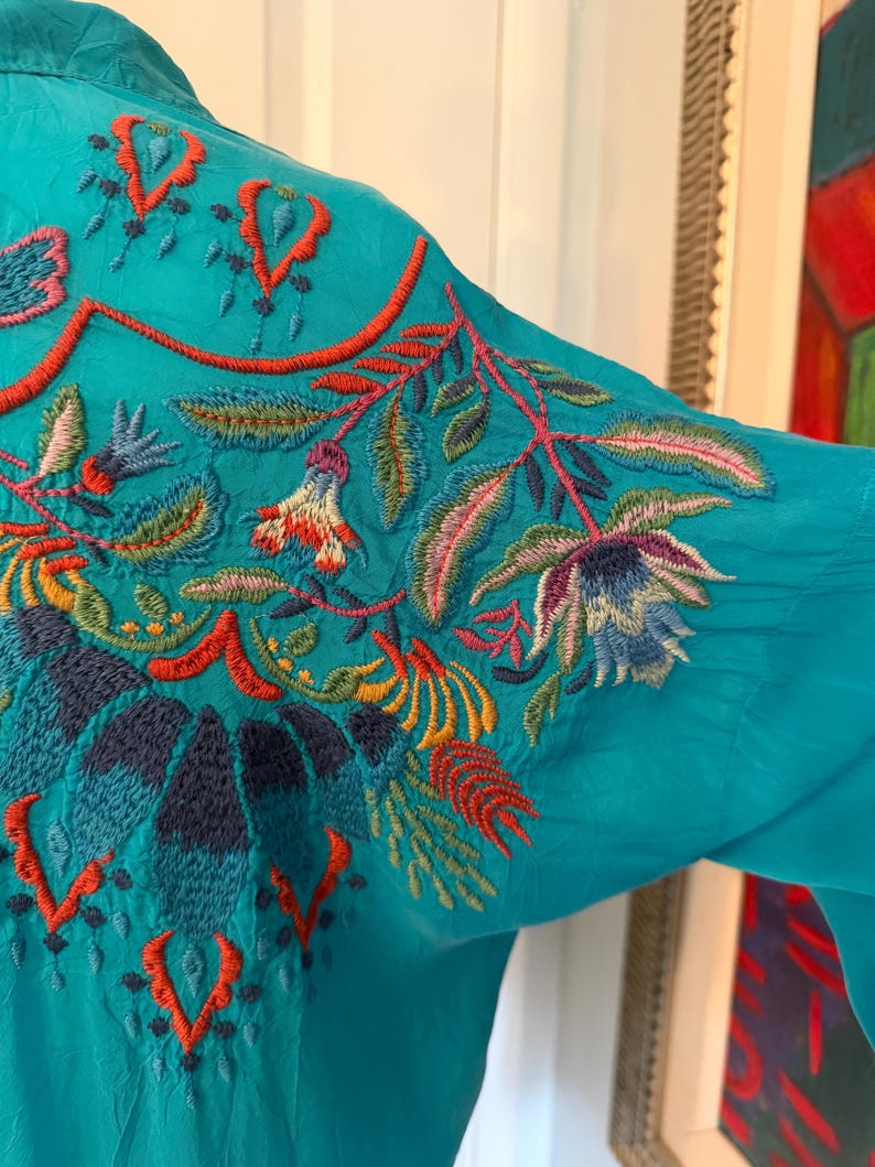May include: Teal-colored blouse with intricate floral embroidery in shades of red, orange, blue, green, and yellow. The embroidery is concentrated on the upper back and shoulder area, creating a vibrant and detailed design. The blouse has long sleeves.