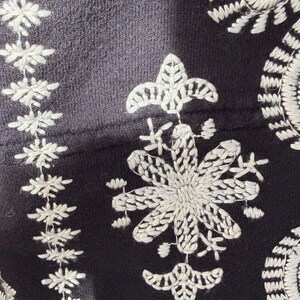 K&ouml;nnte beinhalten: Nahaufnahme eines schwarzen Stoffes mit detaillierter wei&szlig;er Blumenstickerei. Das Design zeigt eine gro&szlig;e, stilisierte Blume und Blattmuster. Die Stickerei ist aufwendig und erzeugt einen strukturierten Effekt auf dem dunklen Hintergrund.