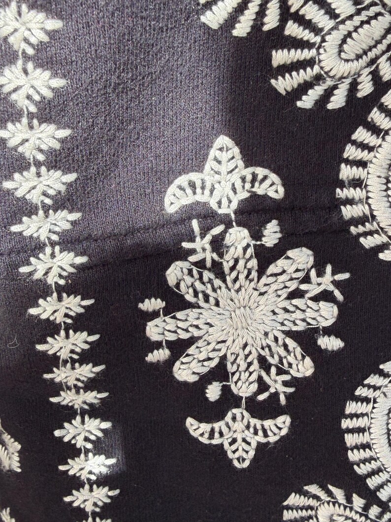 K&ouml;nnte beinhalten: Nahaufnahme eines schwarzen Stoffes mit detaillierter wei&szlig;er Blumenstickerei. Das Design zeigt eine gro&szlig;e, stilisierte Blume und Blattmuster. Die Stickerei ist aufwendig und erzeugt einen strukturierten Effekt auf dem dunklen Hintergrund.
