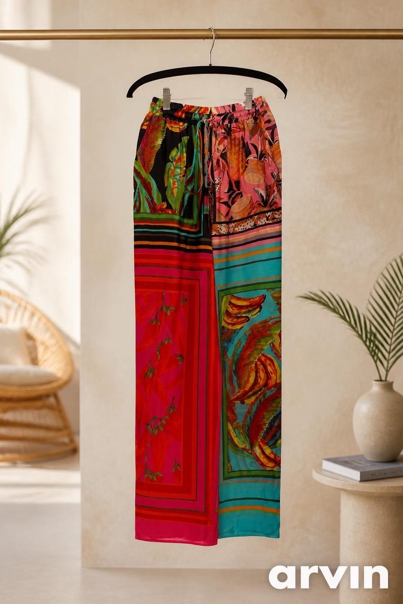 Puede incluir: Pantalones palazzo coloridos y estampados colgados de una percha negra. Los pantalones presentan una mezcla de colores y dise&ntilde;os vibrantes, incluyendo paneles rojos, rosas, turquesas y negros con estampados florales y abstractos. La palabra "arvin" es visible en la parte inferior.