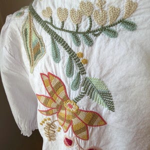 May include: White blouse with detailed floral embroidery in shades of yellow, green, and pink. The embroidery features a large flower, leaves, and heart-shaped accents. The blouse has short sleeves and a round neckline.