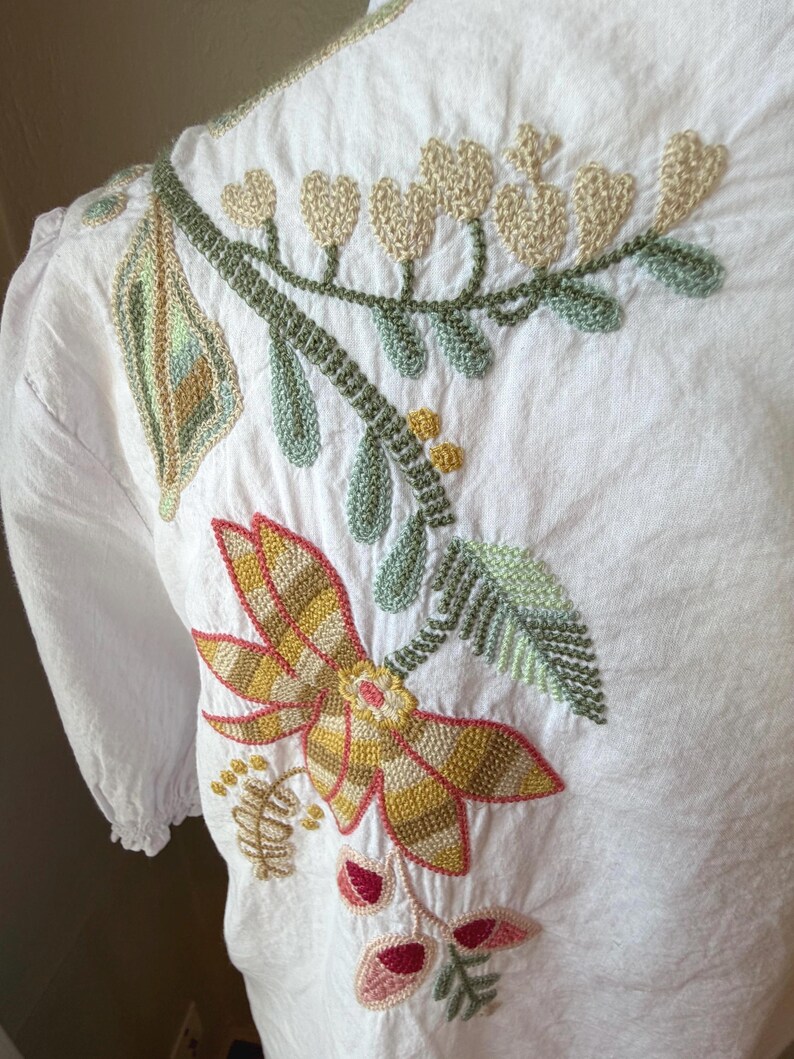 May include: White blouse with detailed floral embroidery in shades of yellow, green, and pink. The embroidery features a large flower, leaves, and heart-shaped accents. The blouse has short sleeves and a round neckline.