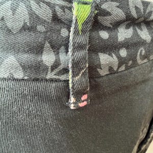 Puede incluir: Un pantal&oacute;n gris carb&oacute;n oscuro con una cintura ancha con un patr&oacute;n floral en negro y gris. Una trabilla &uacute;nica est&aacute; centrada en la cintura, mostrando un peque&ntilde;o acento triangular verde en la parte superior y diminutos puntos rosas abajo.