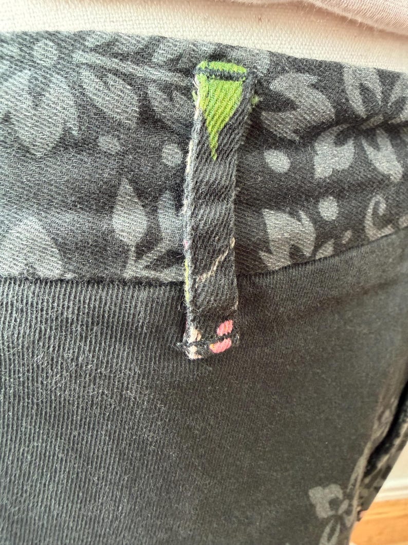 Puede incluir: Un pantal&oacute;n gris carb&oacute;n oscuro con una cintura ancha con un patr&oacute;n floral en negro y gris. Una trabilla &uacute;nica est&aacute; centrada en la cintura, mostrando un peque&ntilde;o acento triangular verde en la parte superior y diminutos puntos rosas abajo.