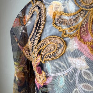 May include: Close-up of a sheer garment with intricate floral and paisley embroidery. The design features a combination of colors, including gold, brown, pink, and green, set against a dark background. The fabric has a delicate, net-like texture.
