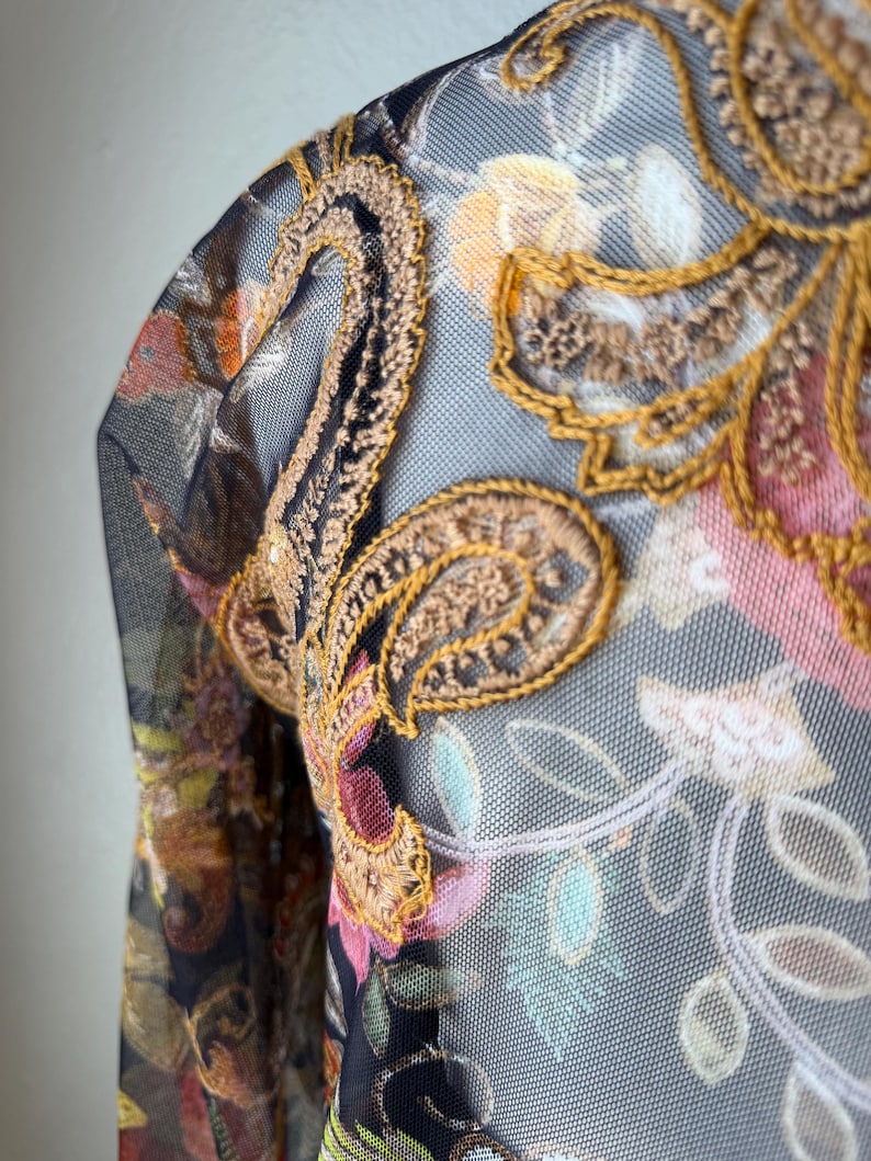 May include: Close-up of a sheer garment with intricate floral and paisley embroidery. The design features a combination of colors, including gold, brown, pink, and green, set against a dark background. The fabric has a delicate, net-like texture.