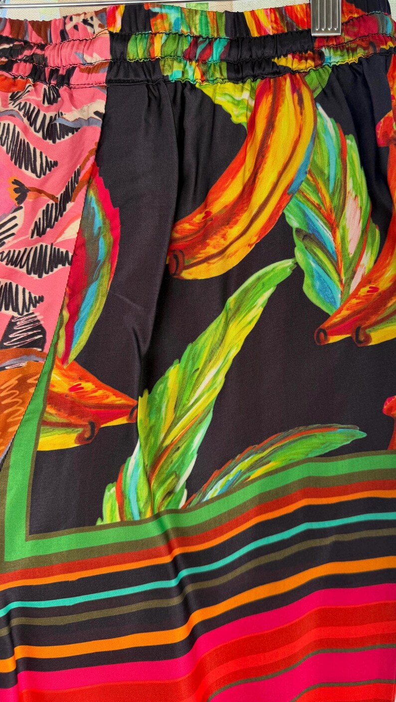 Puede incluir: Primer plano de una tela colorida con estampado tropical. El dise&ntilde;o presenta grandes hojas de pl&aacute;tano estilizadas en tonos amarillos, verdes y rojos sobre un fondo negro. La tela tambi&eacute;n tiene una secci&oacute;n rosa con un patr&oacute;n blanco y un borde a rayas.