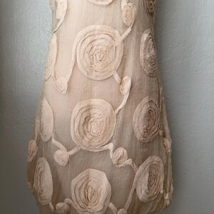May include: A light beige dress with a floral pattern. The dress features large, circular, raised floral appliqu&eacute;s connected by delicate stems. The fabric appears sheer, and the dress is displayed on a white mannequin.