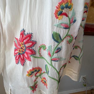 May include: White tunic with colorful floral embroidery. The embroidery features red, pink, yellow, blue, and green flowers and leaves. The tunic has a loose fit and a decorative hemline.