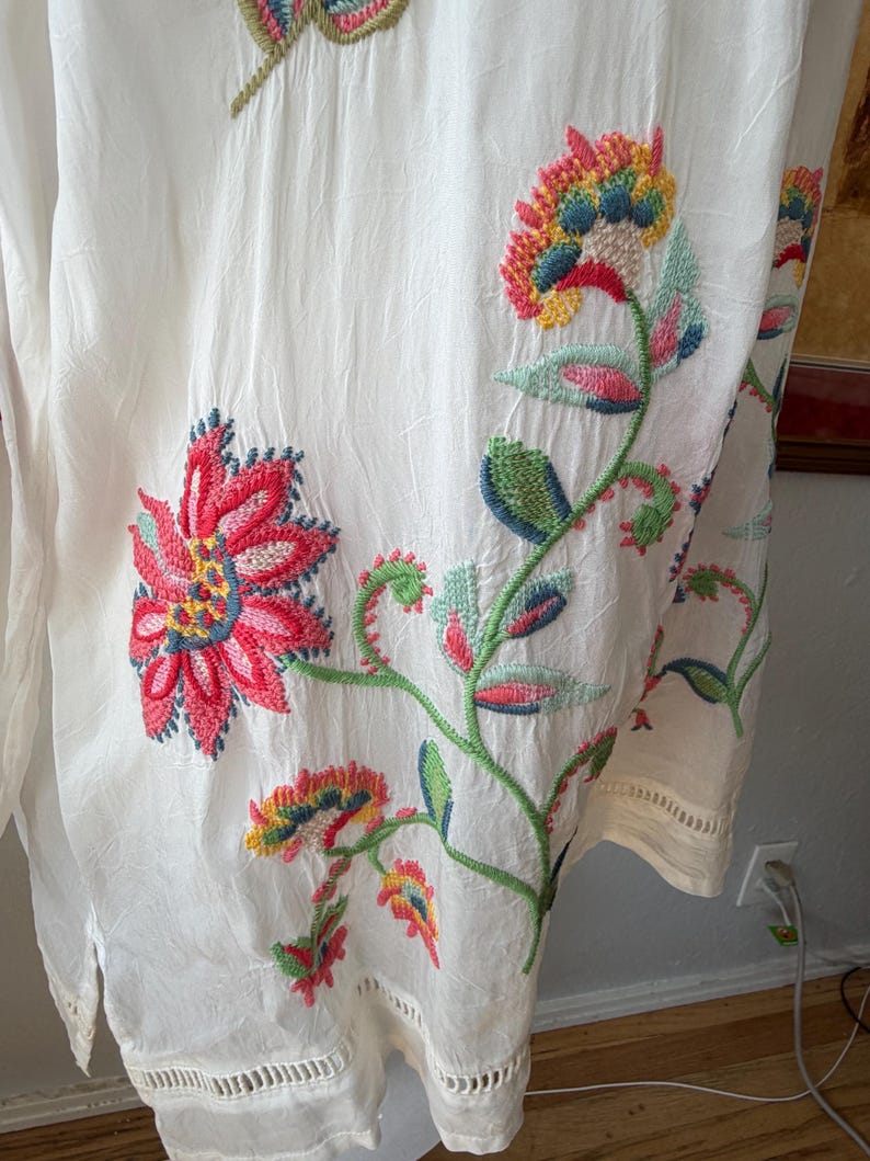 May include: White tunic with colorful floral embroidery. The embroidery features red, pink, yellow, blue, and green flowers and leaves. The tunic has a loose fit and a decorative hemline.